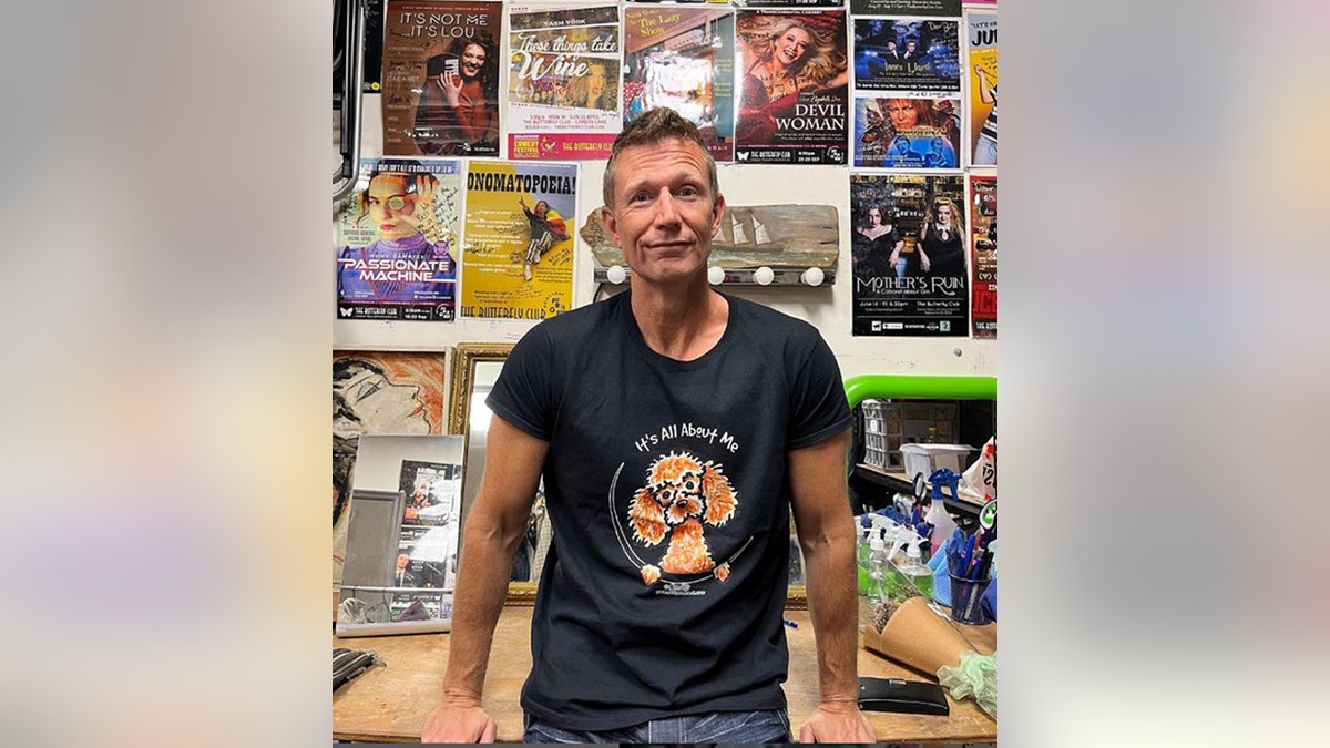 Tony Knight in a graphic shirt soft smiles in front of a wall of posters